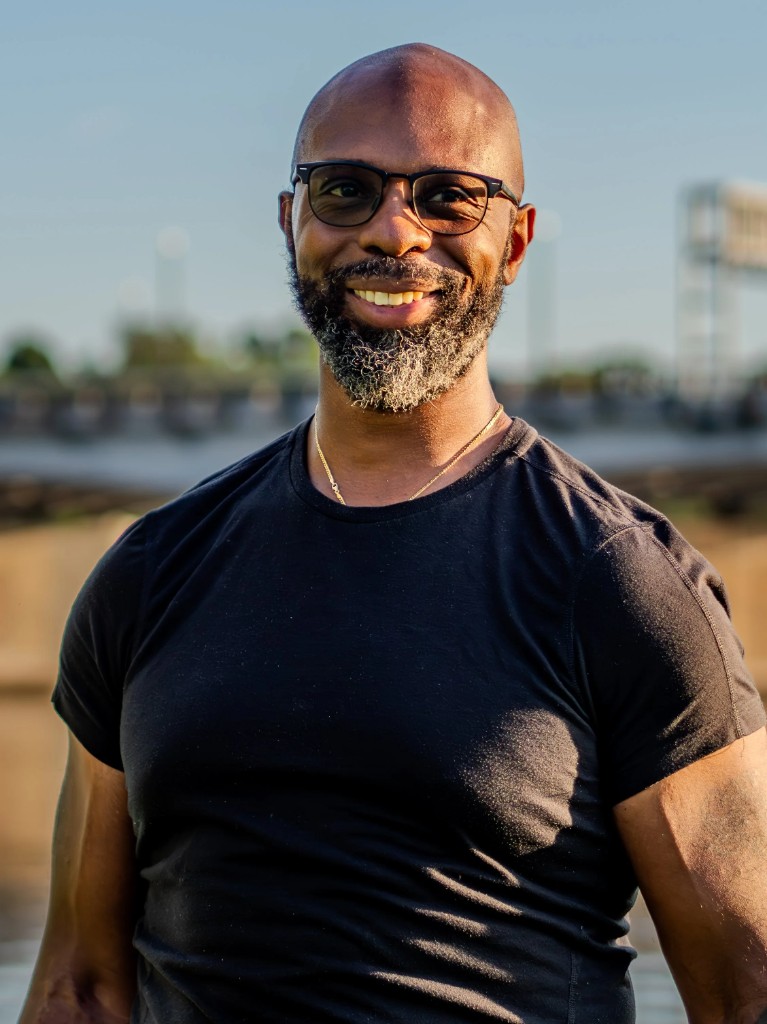 Professional portrait of Coach Adam, smiling, with a salt-and-pepper beard and glasses, wearing a black t-shirt.